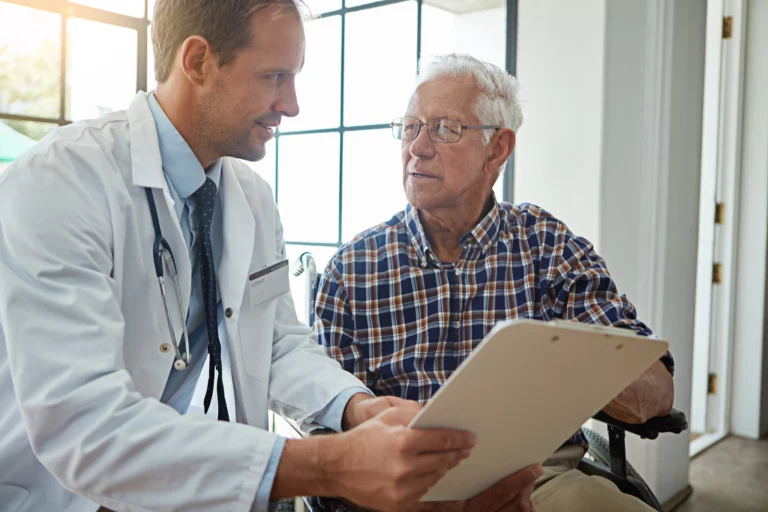 A doctor in a white coat and stethoscope reviews a clipboard with an elderly man in a wheelchair, engaged in a focused conversation.